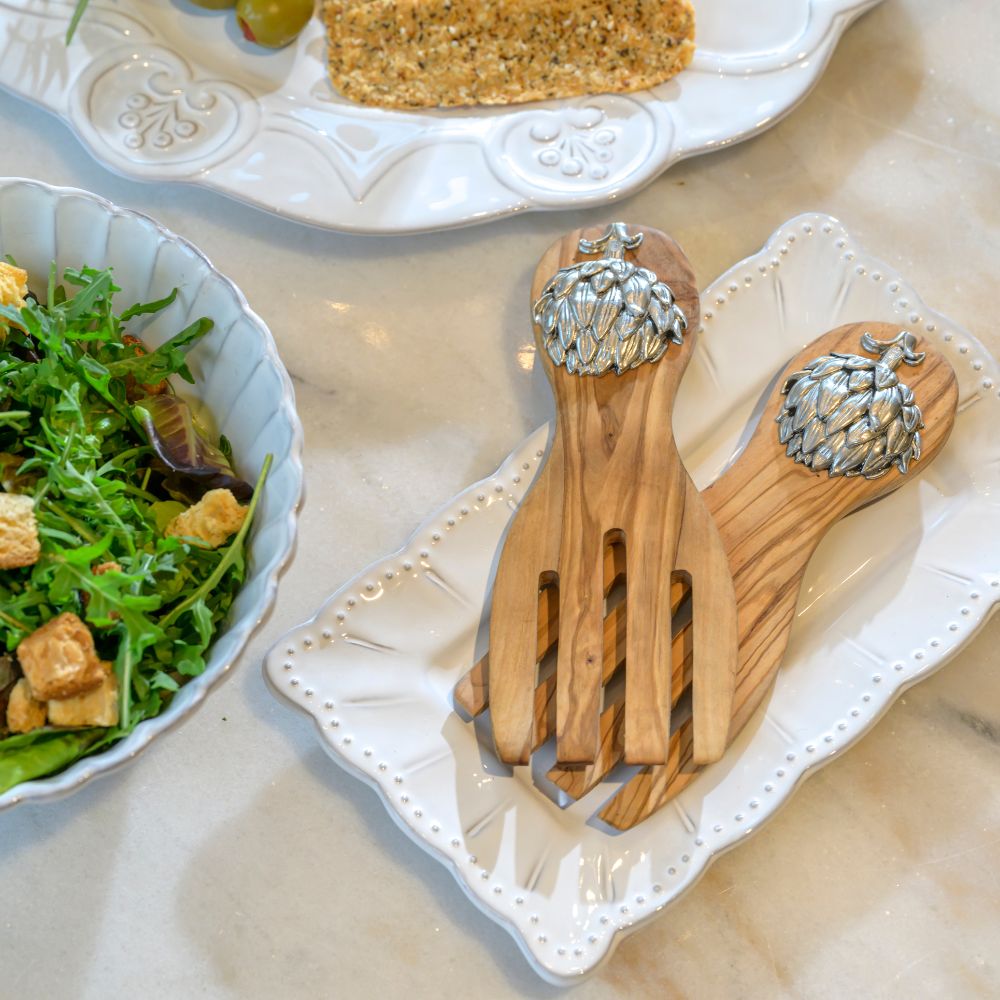 Olive Wood Salad Servers with pewter Artichoke decoration on handles placed on a decorative plate with salad and bread.