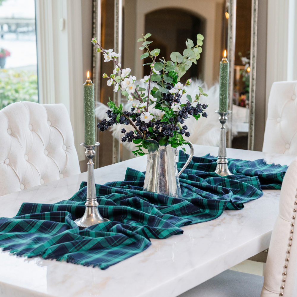 Black watch tartan runner on a white marble table with two Alaria pewter candlesticks and a pewter pitcher with greens and berries