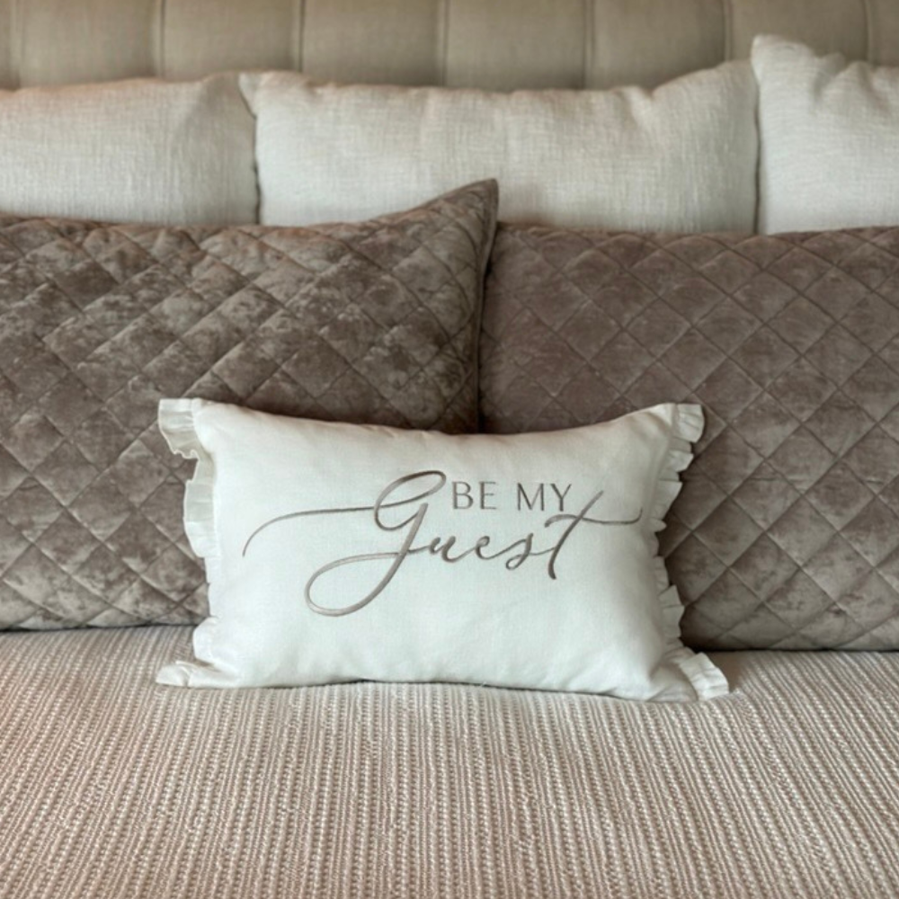 Decorative pillow in 100% linen with 'Be My Guest' embroidered text on a bed with small ruffles