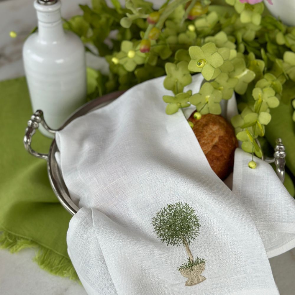 White linen towel with embroidered herb topiary design on a table setting with flowers and a bread roll.