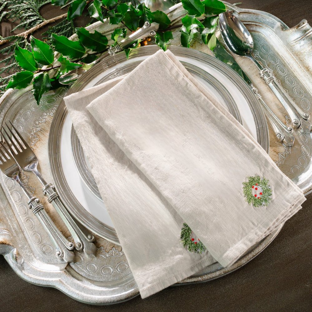 Two folded soft flax linen napkins embroidered in the corner with a mini wreath with red berries on a silver tray with holly