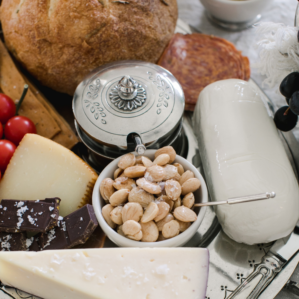 Top view of the Peltro pewter lid condiment dish with spoon on a charcuterie board with nuts, bread, cheese and chocolate