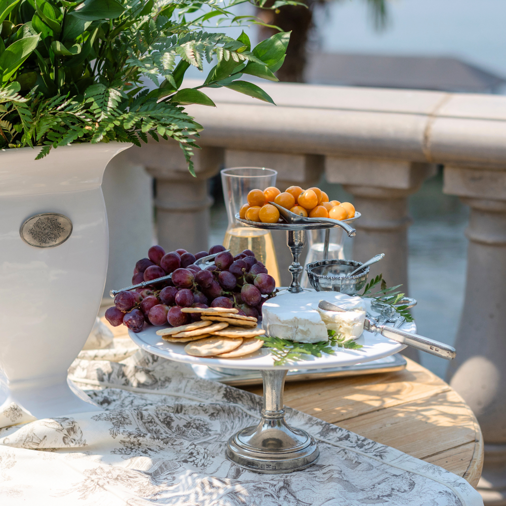 Tuscan cake stand on a table overlooking the water set with cheese, crackers and grapes.