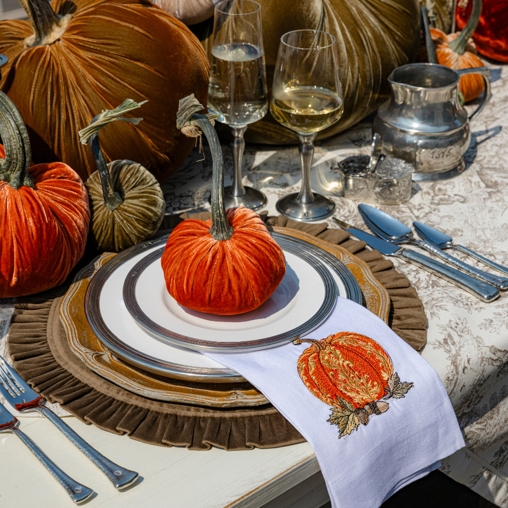 A baroque embroidered pumpkin in orange on a white linen towel on a brown velvet round ruffle placemat with velvet pumpkins on a table