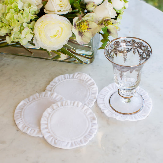 A set of white velvet coasters with a frilled edge displayed on a table, with one coaster slightly lifted to show the texture.