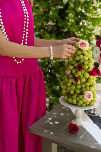 Person in a pink dress and pearl bracelet arranging a decorative fruit tower with grapes and roses on a table.