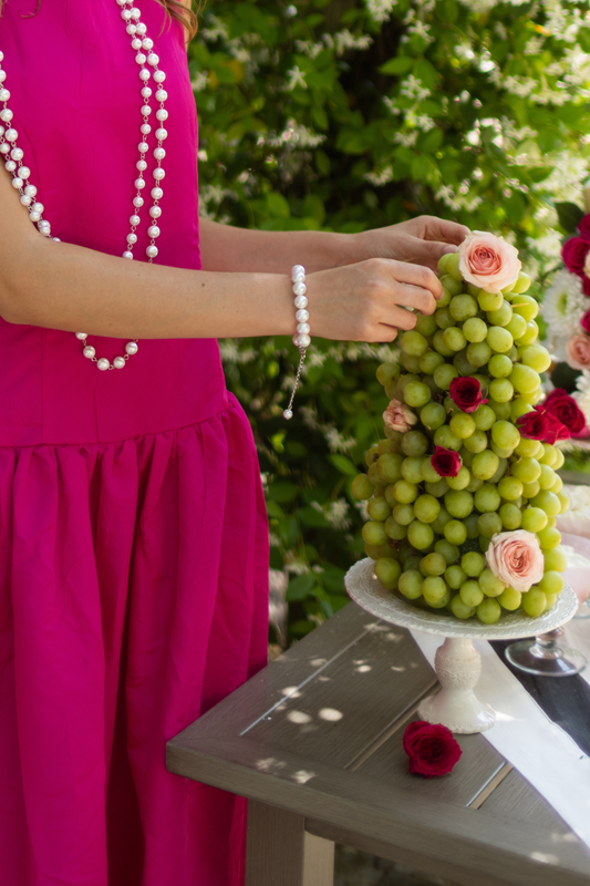 Person in a pink dress and pearl bracelet arranging a decorative fruit tower with grapes and roses on a table.