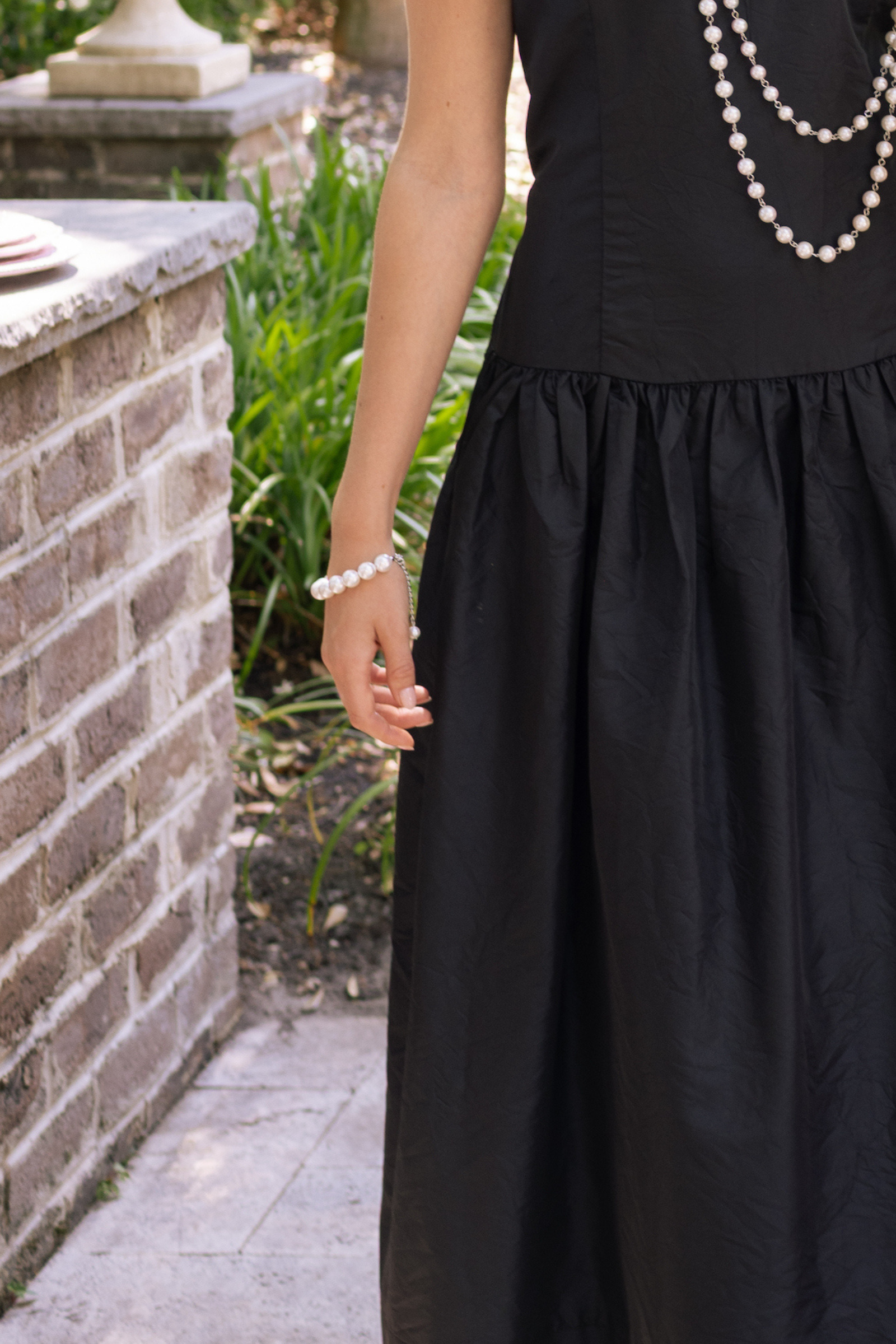 Person wearing a black dress with a pearl necklace and bracelet in an outdoor setting.