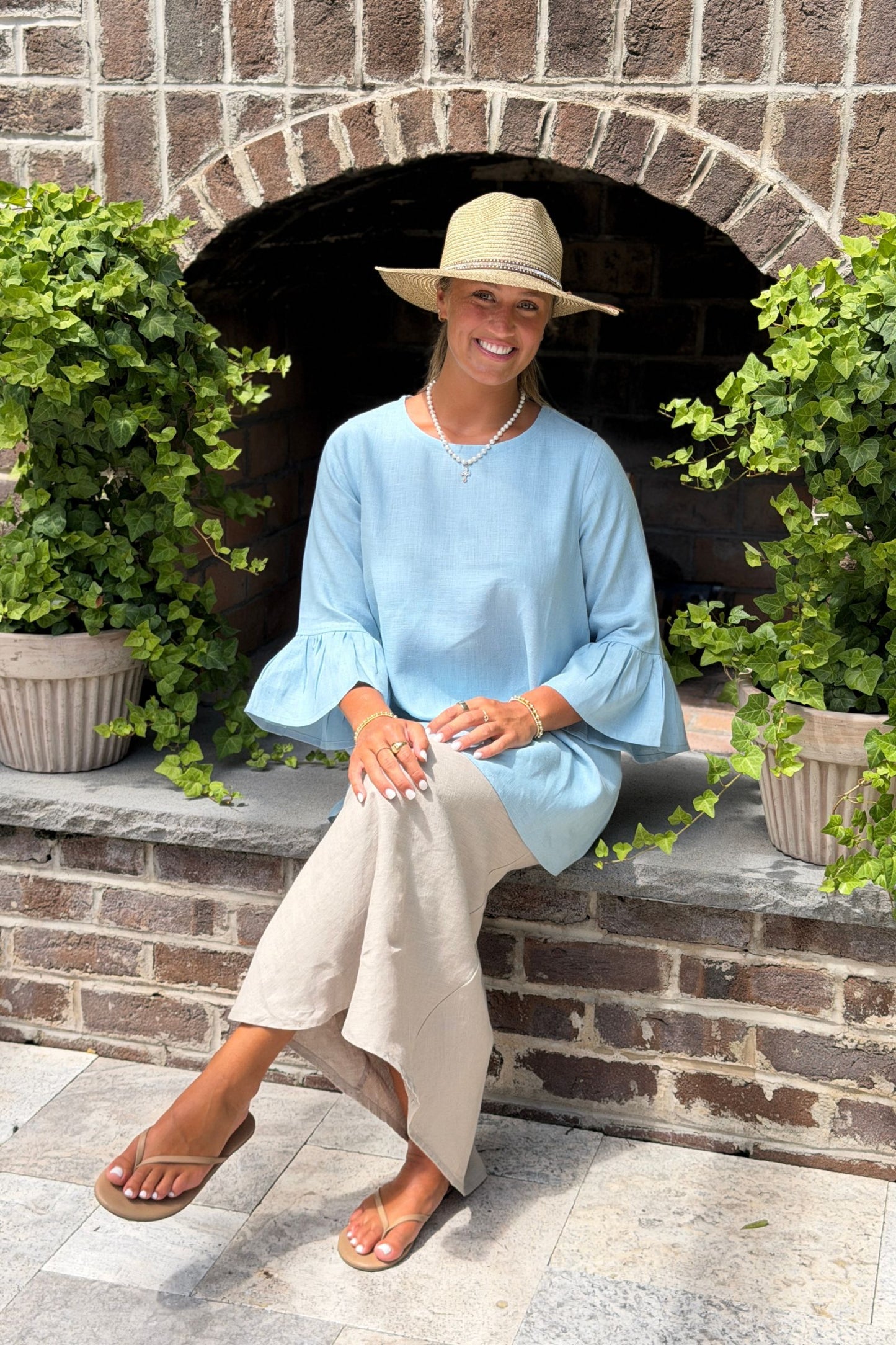 A woman wearing a light blue linen Bella blouse with bell sleeves and a beige wide-brim hat sits on a stone hearth in front of a brick outdoor fireplace with potted green plants.