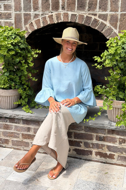 A woman wearing a light blue linen Bella blouse with bell sleeves and a beige wide-brim hat sits on a stone hearth in front of a brick outdoor fireplace with potted green plants.