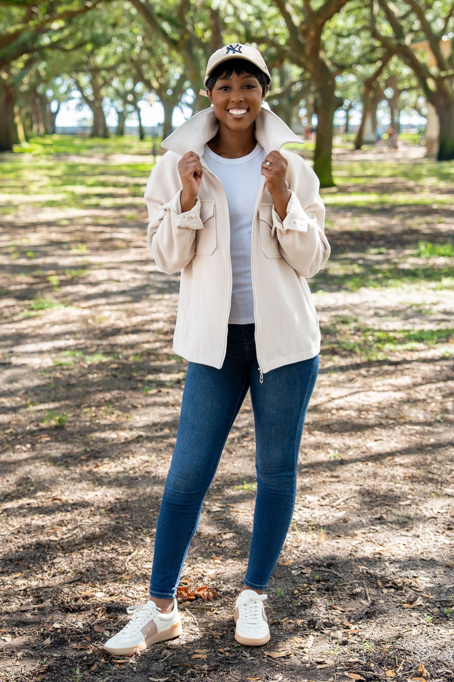 Woman standing outdoors in a park wearing a Wool Blend latte colored Alex Jacket, white shirt, blue jeans, and white sneakers.