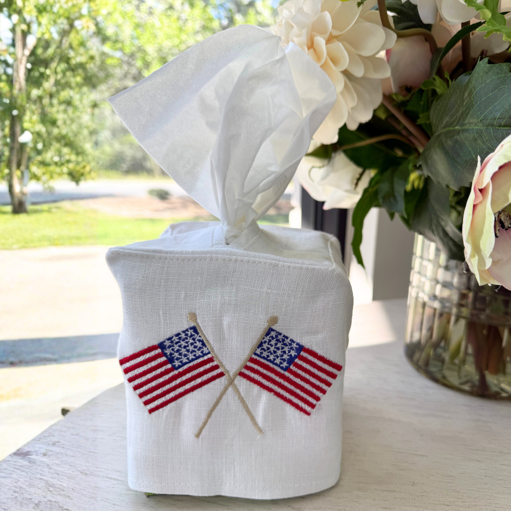 A white linen tissue box holder embroidered with American flags sitting on a table near a window with flowers in the background