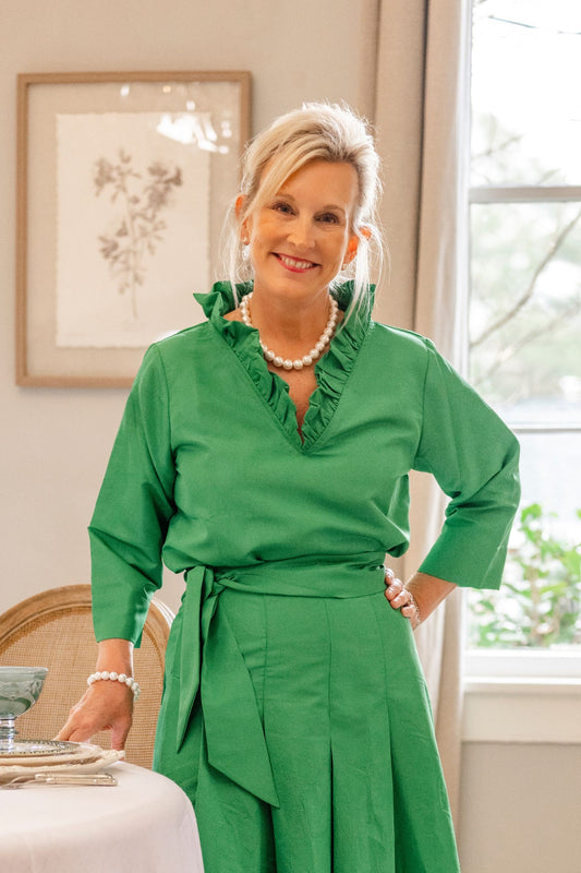 A woman in a green Darcy Taffeta Top standing in a room with a window and a framed picture.