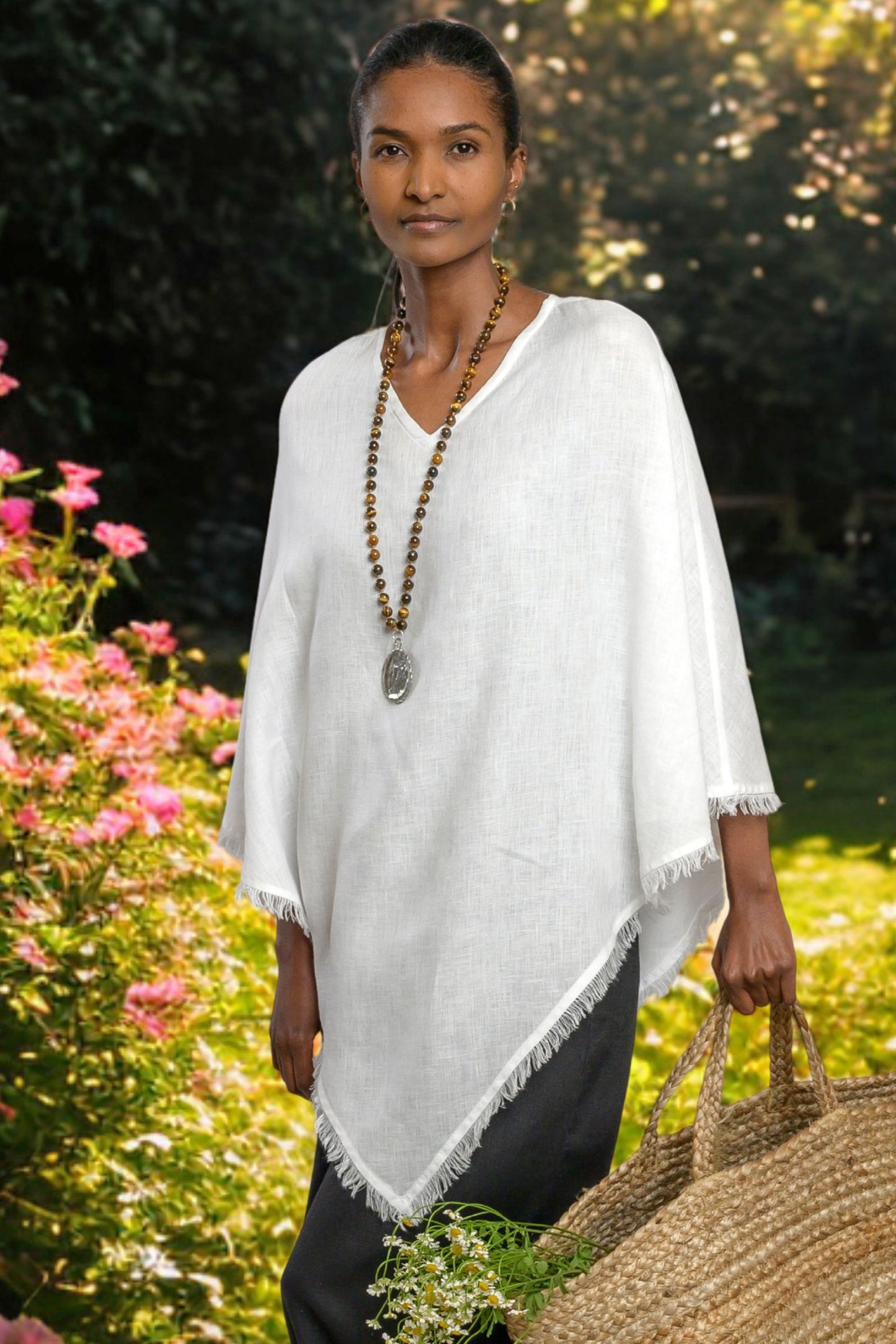 Woman wearing a white linen poncho in a garden setting with a brown necklace. --Arte Italica and Crown Linen Designs