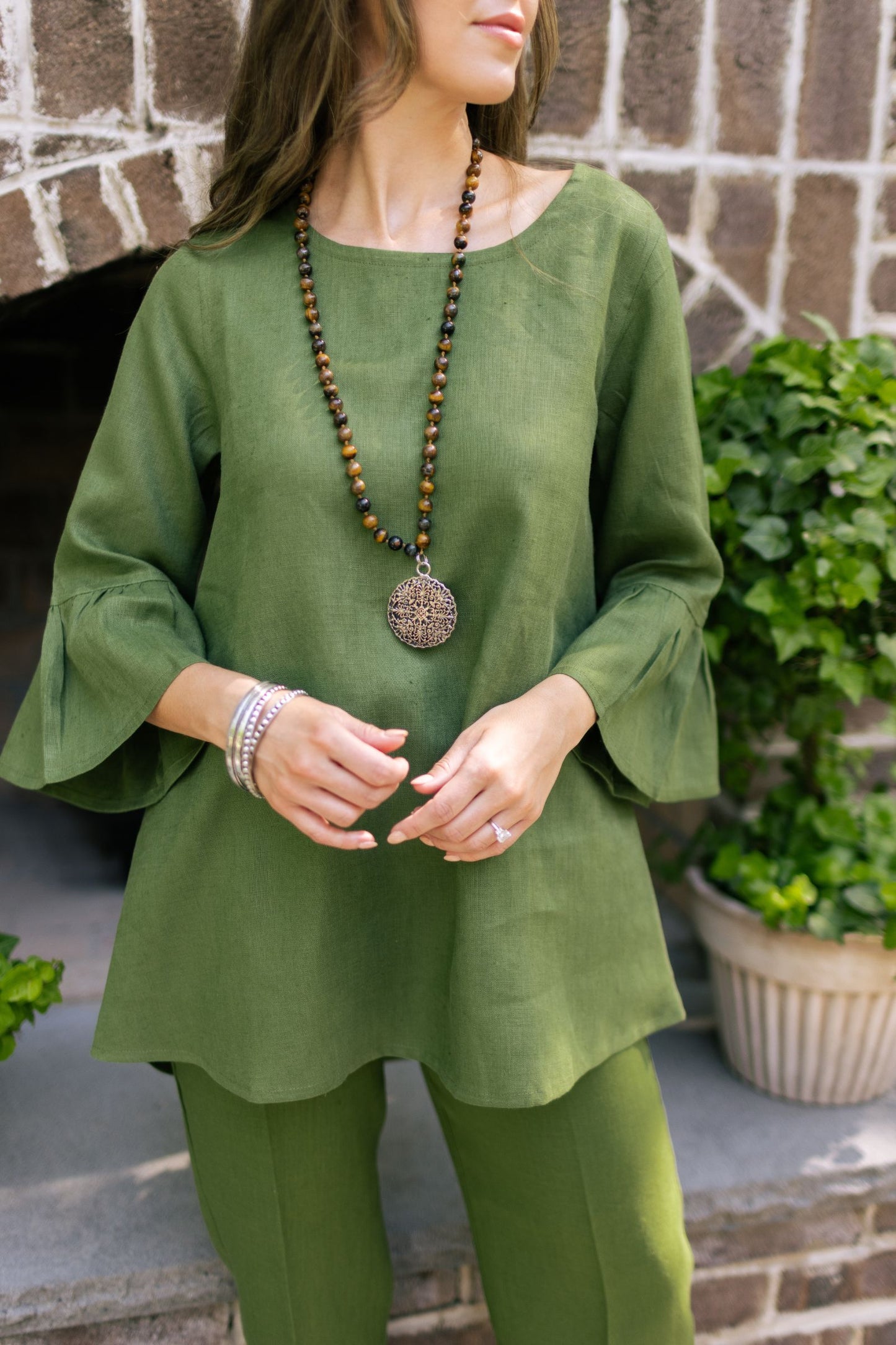Woman posing wearing a green linen top with 3/4 sleeves with ruffles and green linen capri pants with a brown necklace and pewter bangles. --Arte Italica and Crown Linen Designs