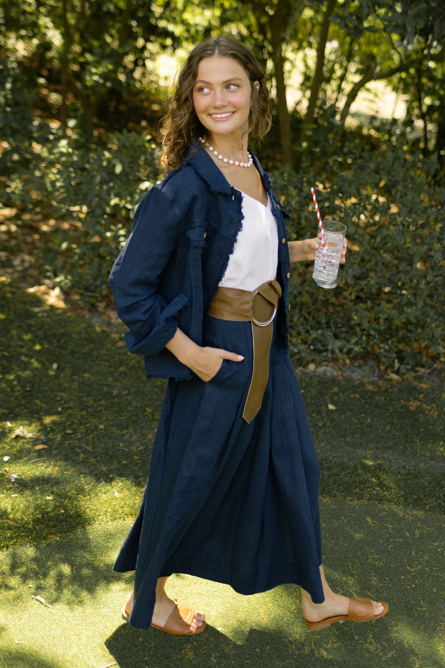 Woman in a navy Jessie jacket and Lizzy linen skirt outfit with a white linen cami top and brown belt, holding a drink, standing outdoors. --Arte Italica and Crown Linen Designs