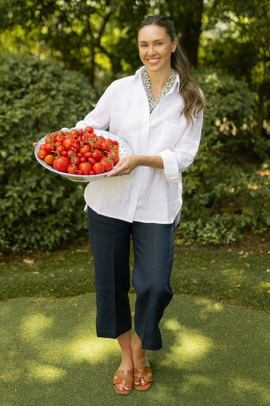 Woman wearing white linen Belmont button up top with Navy Classic Capri pants paired with cluster beaded necklace while posing outside with bowl of tomatoes. --Arte Italica and Crown Linen Designs