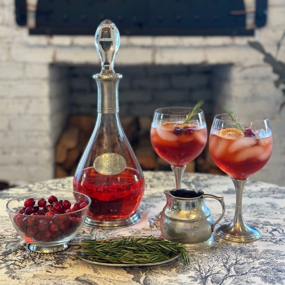 Red cocktail with ice and garnish in two Verona glasses, accompanied by a Damasco Elegante decanter and bowl of cranberries on a toile tablecloth.