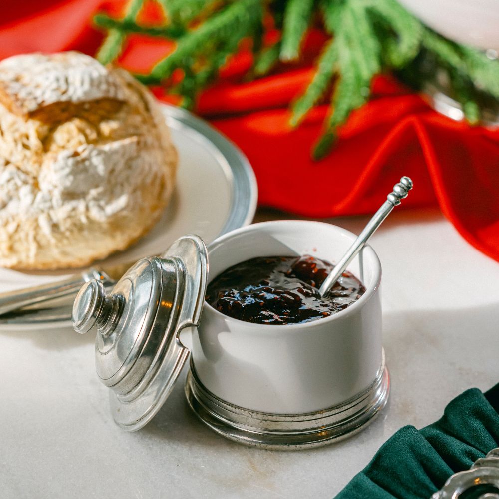 White Tuscan ceramic jar of jam with a silver lid and spoon, next to a scone on a marble surface with a red napkin and greenery in the background.