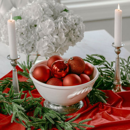 Decorative table setting with red ornaments in a bowl, candles, and greenery.