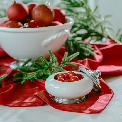 Decorative setting with red ornaments, greenery, and a white ceramic Tuscan Keepsake Box with candy on a red Table square.
