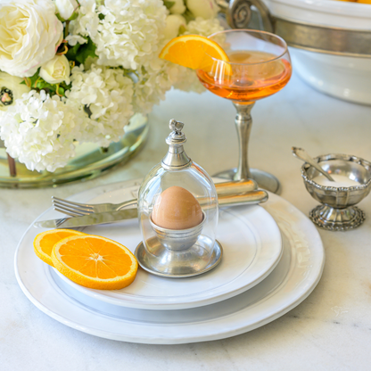 An egg cup with a glass cloche topped with a chick design, placed on a Firenze white plate next to a fork and knife, with orange slices and a vase of flowers in the background.