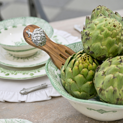 Artichokes in a Green Burano bowl with a wooden artichoke Salad Server on a table with plates in the background