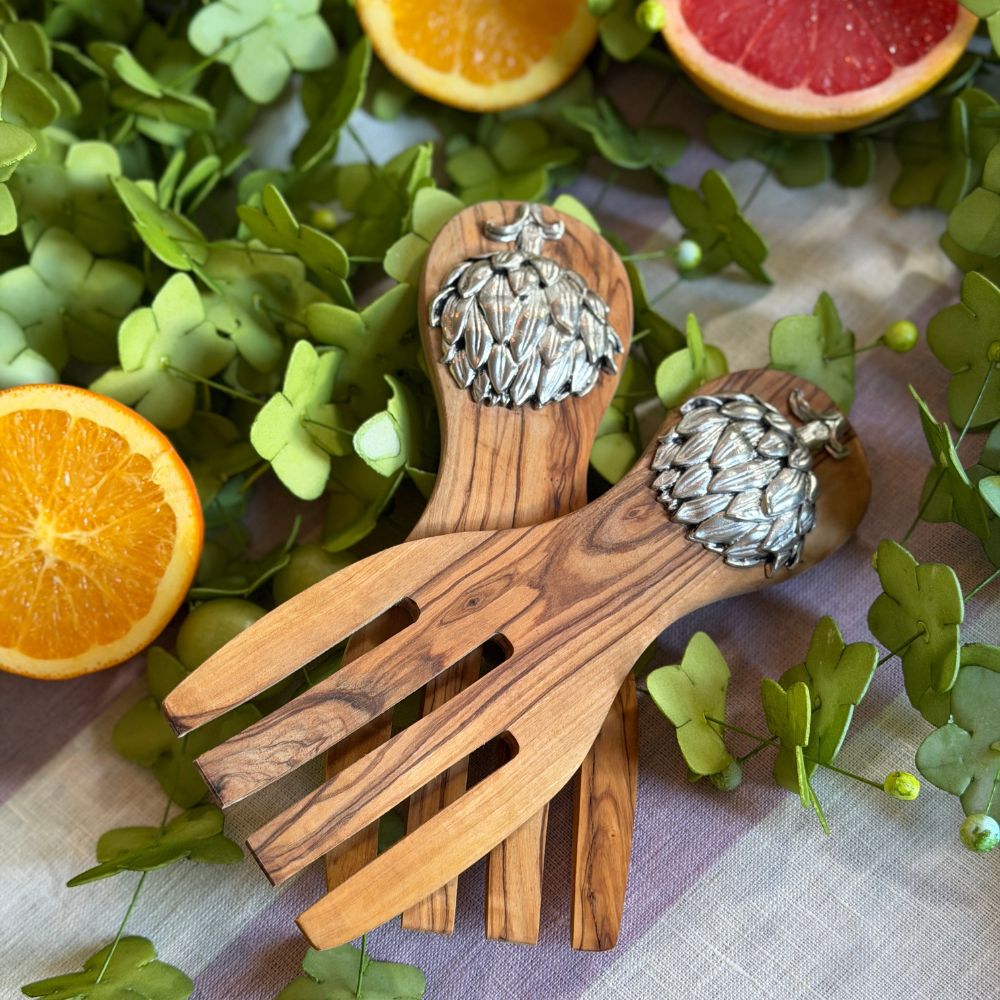 Two wooden salad servers with artichoke pewter decoration on the handles, arranged on a tablecloth and surrounded by green leaves and sliced citrus fruits.