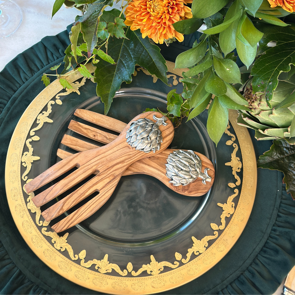 Wooden salad servers with pewter artichoke emblems on a decorative Vetro Gold plate with floral and greenery elements.