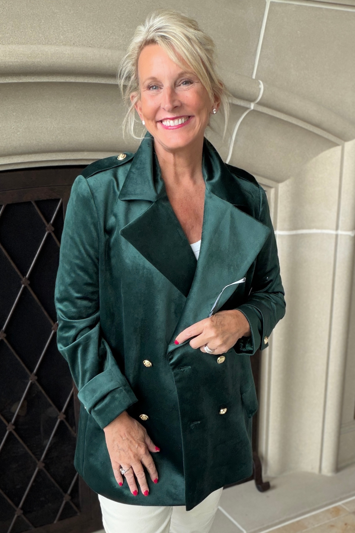 Woman smiling in front of a fireplace wearing the Crown Linen Avery Luxury Velvet Blazer in forest green with gold buttons and white trousers