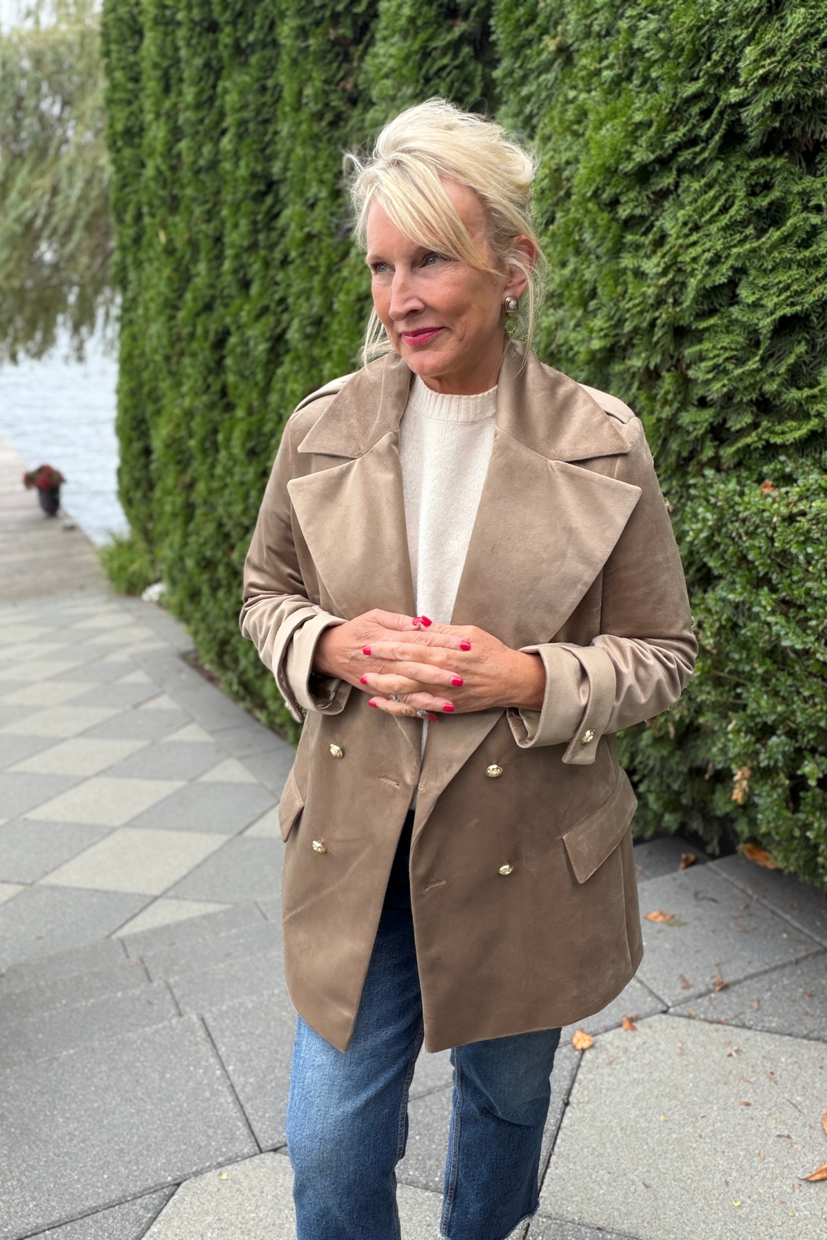 Avery Luxury Velvet Blazer in mocha with gold buttons, styled over a white turtleneck and jeans on an outdoor stone walkway beside a lush green hedge and waterfront