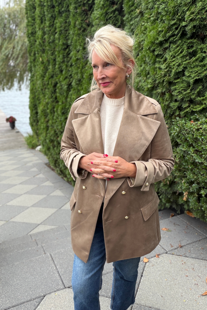 Avery Luxury Velvet Blazer in mocha with gold buttons, styled over a white turtleneck and jeans on an outdoor stone walkway beside a lush green hedge and waterfront