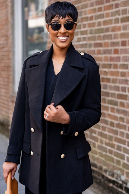 Woman wearing a black Avery wool blend coat with gold buttons against a brick wall.