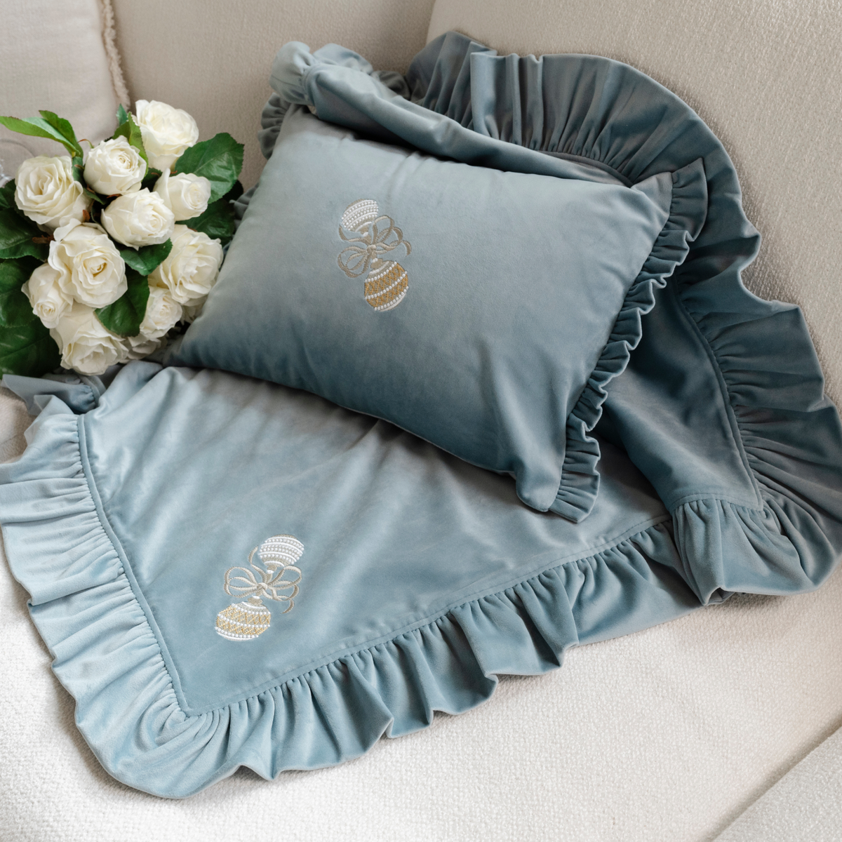 A light blue velvet decor pillow and a small throw embroidered with a baby rattle displayed on a couch with white roses