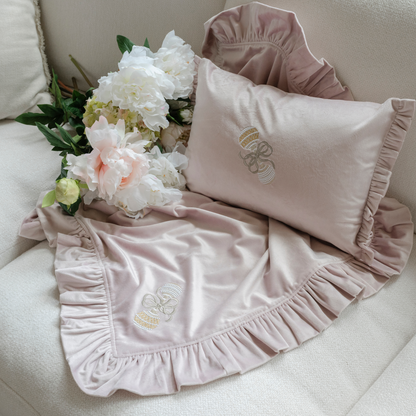A pink velvet decor pillow and a small throw embroidered with a baby rattle displayed on a couch with while flowers