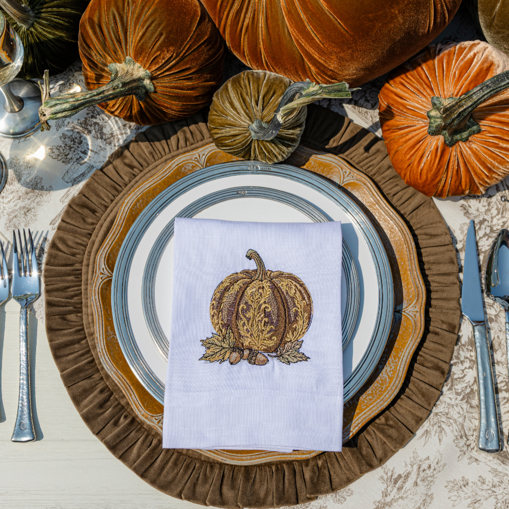 A baroque embroidered pumpkin in brown on a white linen towel on a brown round ruffle placemat with Tuscan with velvet pumpkins on a table