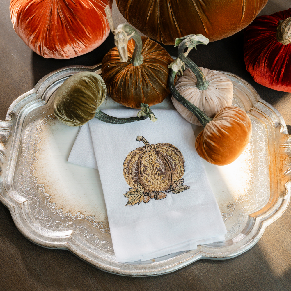 A baroque embroidered pumpkin in brown on a white linen towel on a silver tray with velvet pumpkins on a table