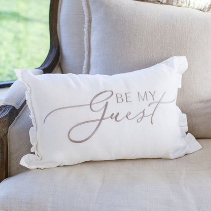 A white decorative pillow with the text 'BE MY GUEST' embroidered on it, displayed on a neutral toned sofa.