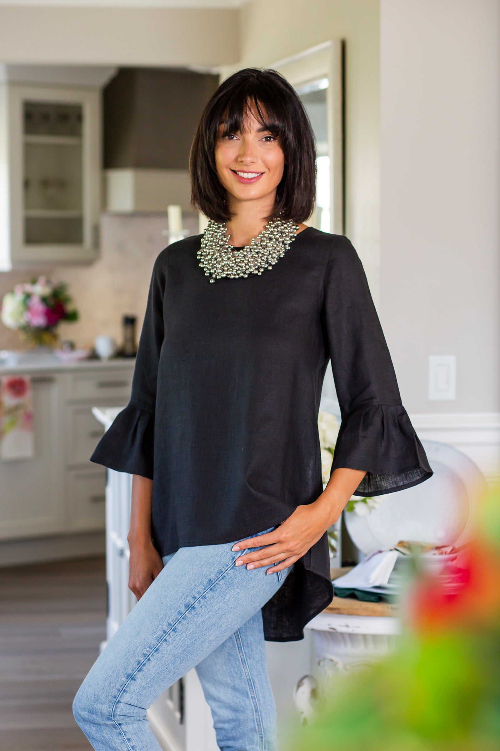 Woman wearing black linen Bella top with 3/4 bell ruffle sleeves and jeans, with a silver beaded cluster necklace posing in a kitchen. --Arte Italica and Crown Linen Designs