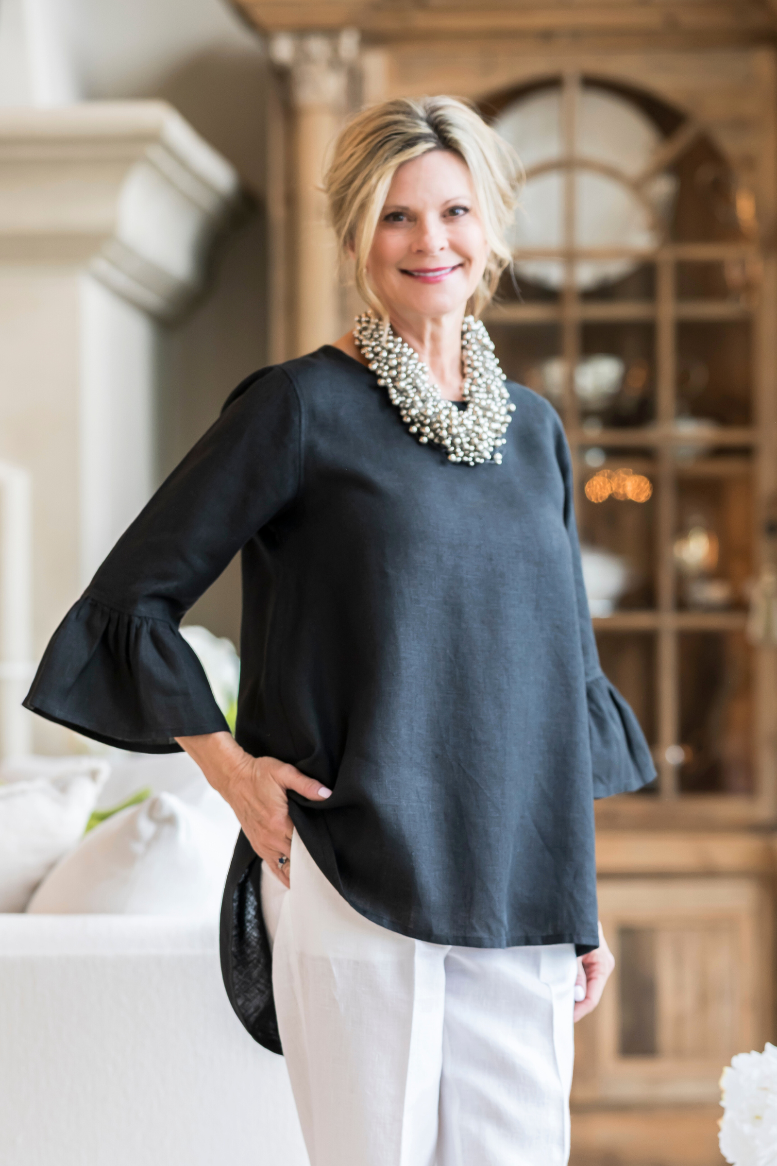 Woman wearing a black linen Bella top with 3/4 sleeves with ruffles and white linen pants with a clustered silver beaded necklace in a living room. --Arte Italica and Crown Linen Designs