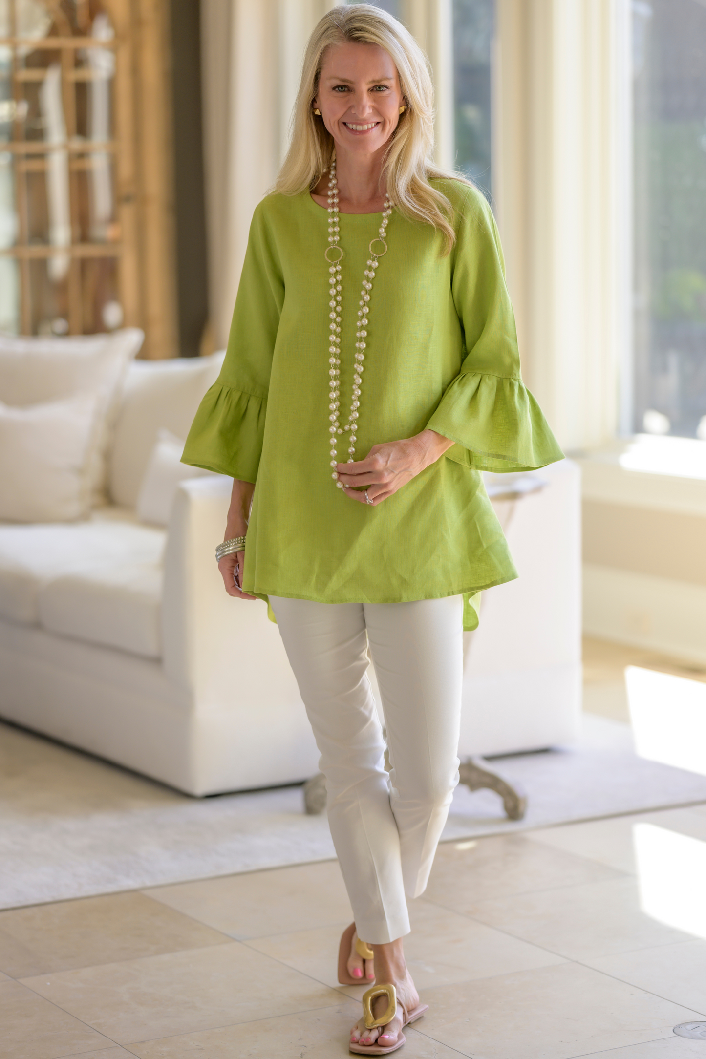 Woman wearing a citron green linen Bella top with 3/4 ruffle sleeves with white pants and a pearl necklace standing in a living room. --Arte Italica and Crown Linen Designs