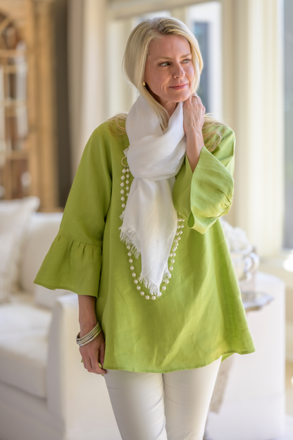 A woman wearing a white fringe Frankie linen scarf, citron green Bella linen top with 3/4 sleeves with ruffles and white pants with a pearl necklace and pewter bangles in a living room. --Arte Italica and Crown Linen Designs