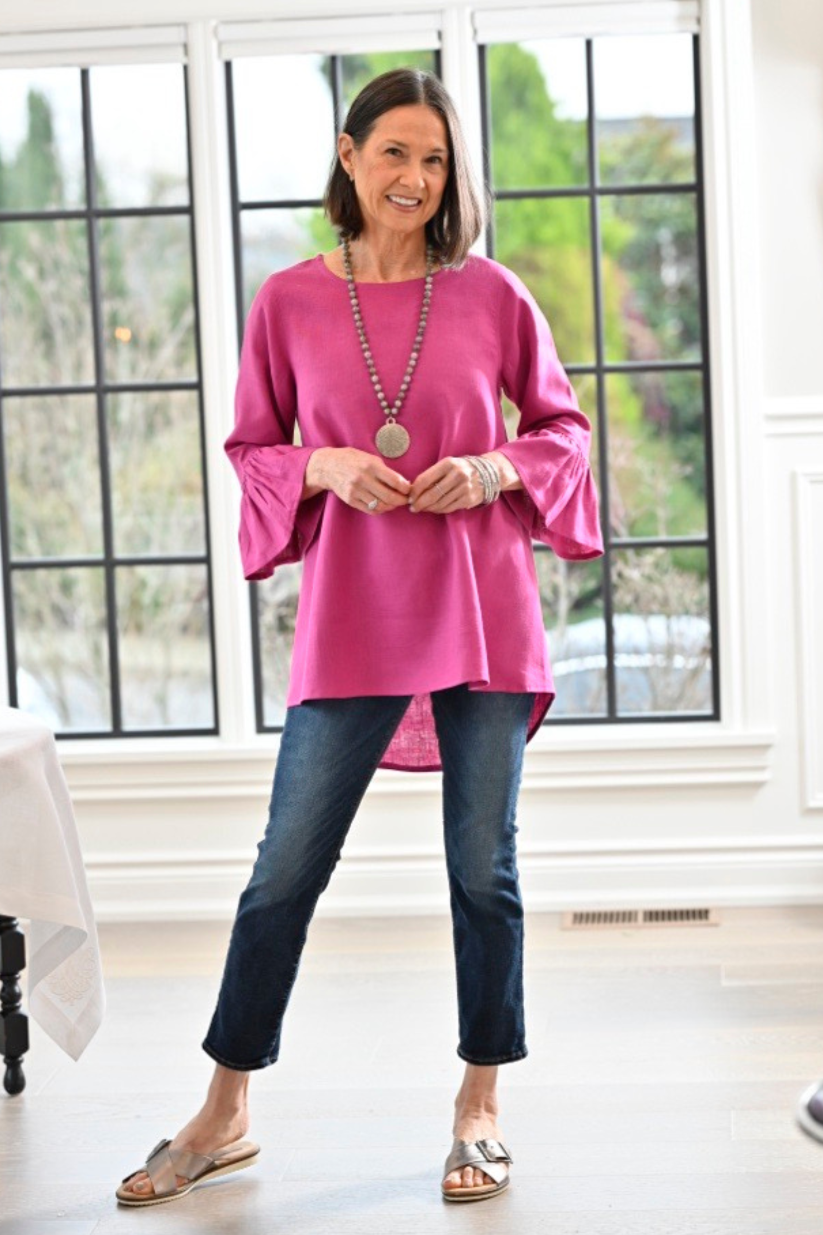 Woman wearing a magenta linen Bella top with 3/4 sleeve with ruffles and jeans with a gray necklace posing by windows. --Arte Italica and Crown Linen Designs