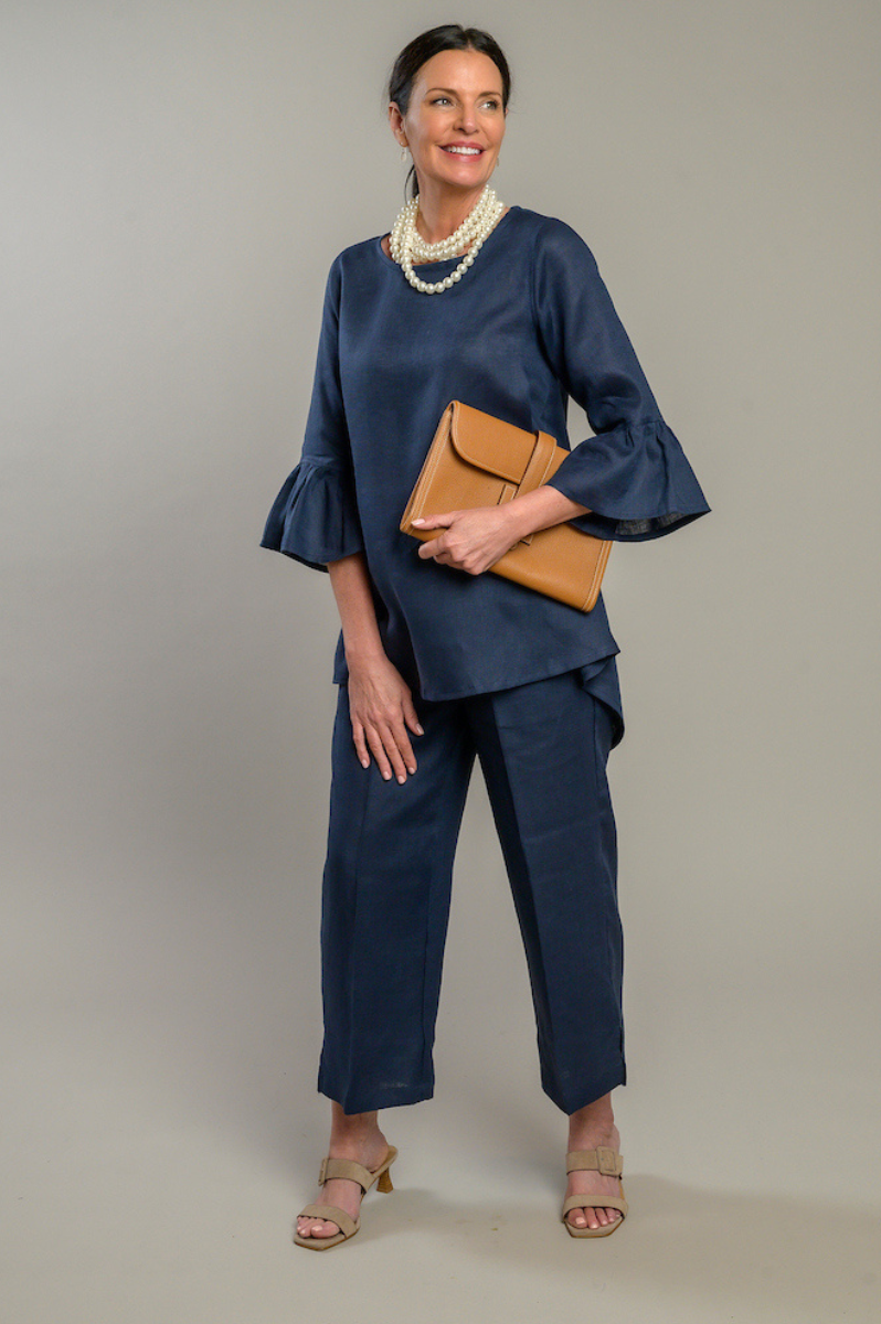Woman wearing a navy Bella linen top with 3/4 sleeves with ruffles and navy linen capri pants pearl necklace holding a ran clutch. --Arte Italica and Crown Linen Designs
