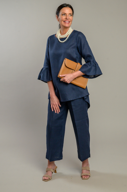 Woman wearing a navy Bella linen top with 3/4 sleeves with ruffles and navy linen capri pants pearl necklace holding a ran clutch. --Arte Italica and Crown Linen Designs