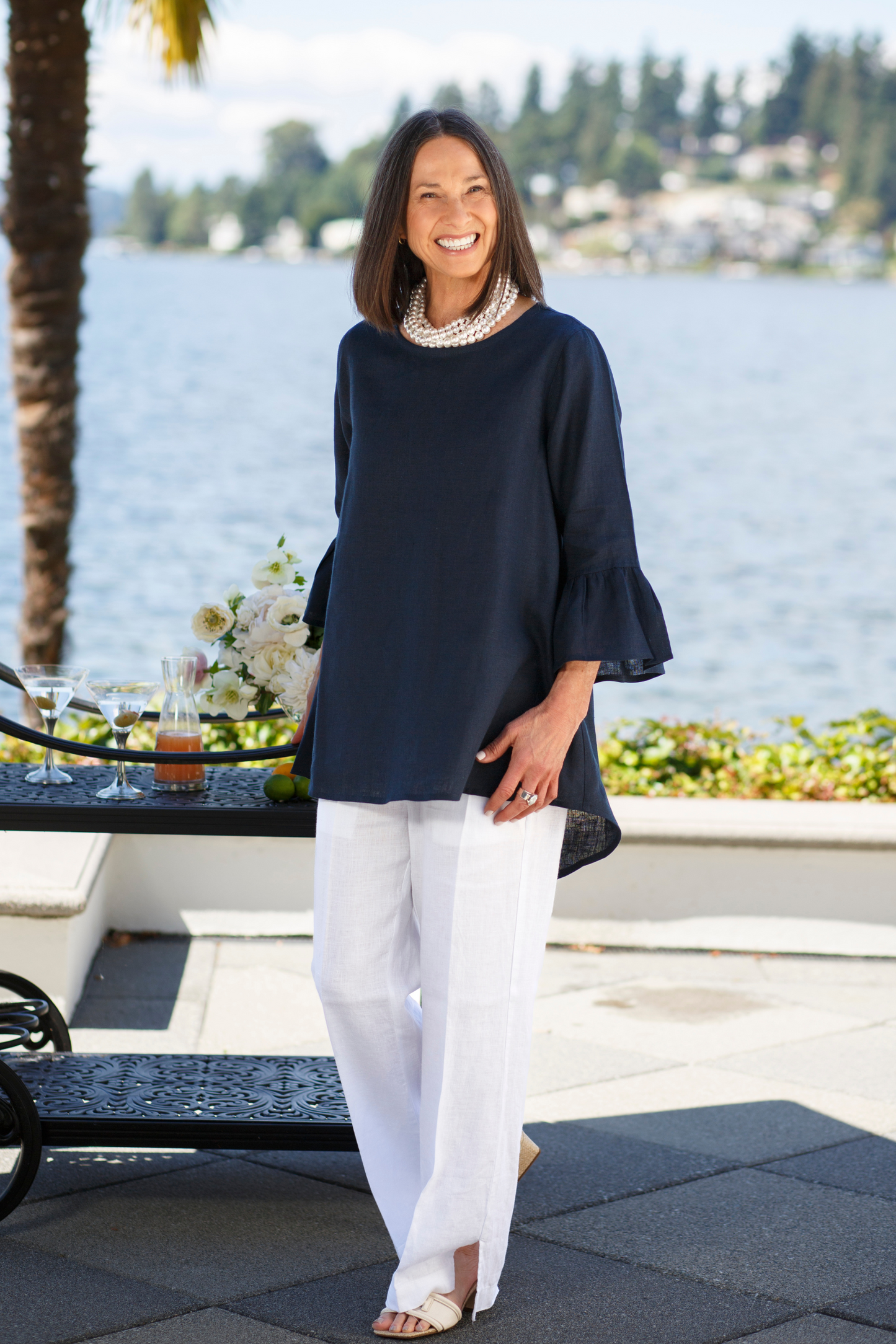 Woman wearing a navy linen Bella top with 3/4 sleeves with ruffles and white linen pants with a pearl stacked necklace posing outside by water. --Arte Italica and Crown Linen Designs