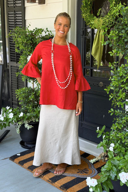 A woman stands on a front porch wearing a red linen Bella top and a long soft flax linen Cindy skirt, accessorized with a long pearl necklace and sandals. The porch is decorated with green plants and a black door.