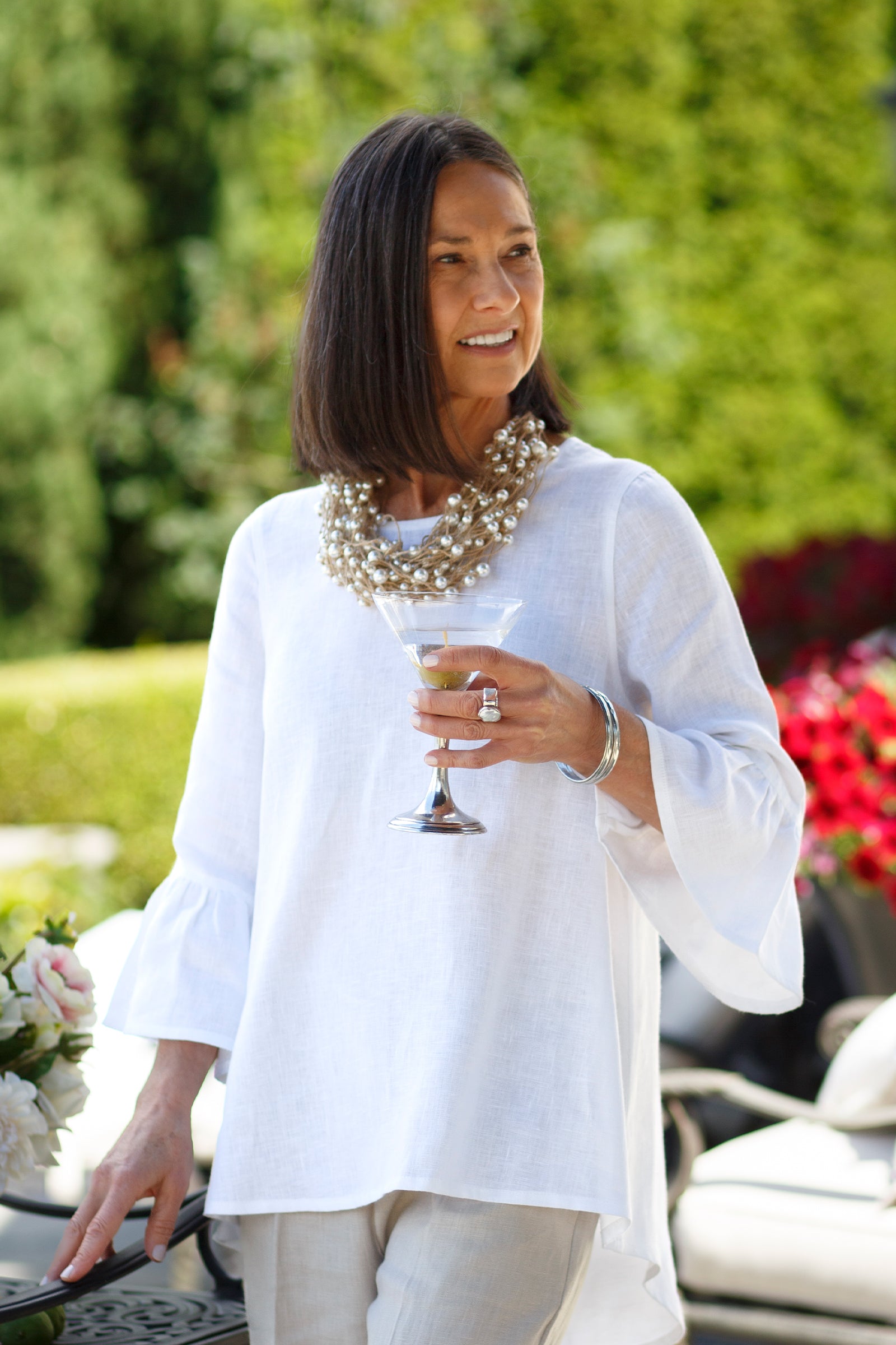 Woman wearing a white Bella linen top with 3/4 sleeves and ruffles with flax colored linen pants with a pearl clustered necklace and pewter bangles holding a martini glass outside. --Arte Italica and Crown Linen Designs