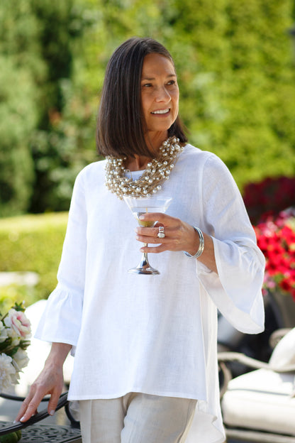 Woman wearing a white Bella linen top with 3/4 sleeves and ruffles with flax colored linen pants with a pearl clustered necklace and pewter bangles holding a martini glass outside. --Arte Italica and Crown Linen Designs