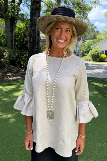 A woman wearing a soft Flax Bella linen blouse with flared three-quarter sleeves, a wide-brimmed straw hat with a black ribbon, and a long pearl necklace, standing outdoors on a green lawn.